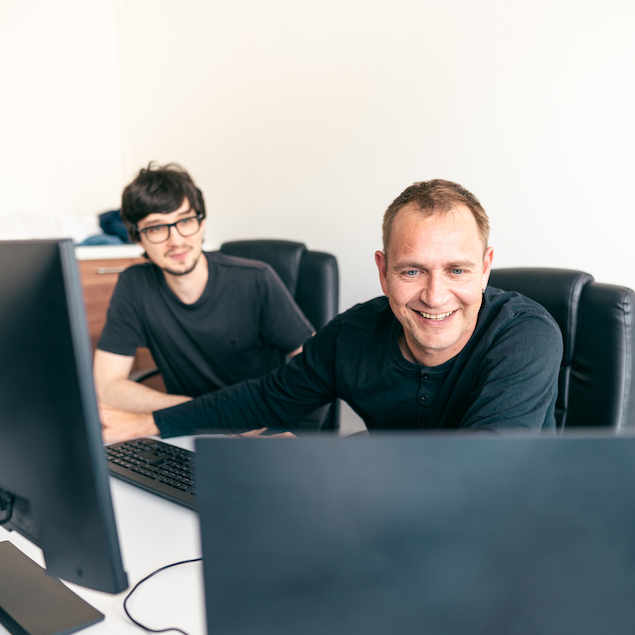 Zwei Lorent IT Mitarbeiter sitzen vor zwei Monitoren, einer lacht